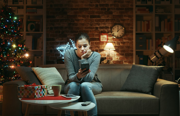 young woman sitting at home during the holidays looking lonely, looking down at smart phone while on couch at home