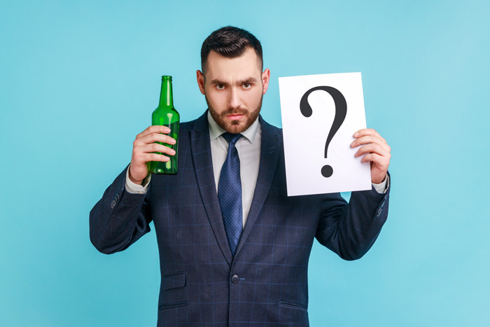 handsome and serious looking businessman holding a green glass beer bottle and a white square sign with a black question mark on it against a sky blue background - alcohol addiction